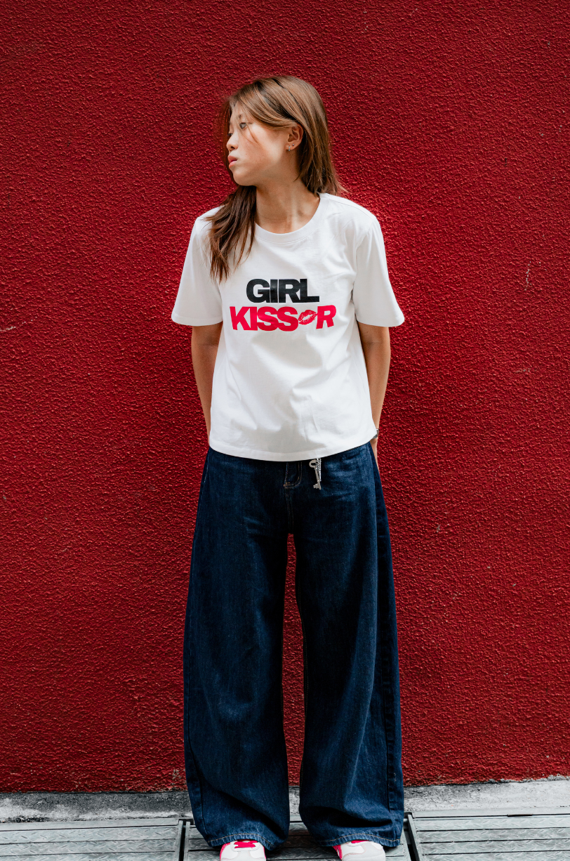 Person wearing a white t-shirt with 'GIRL KISSES' text against a red wall